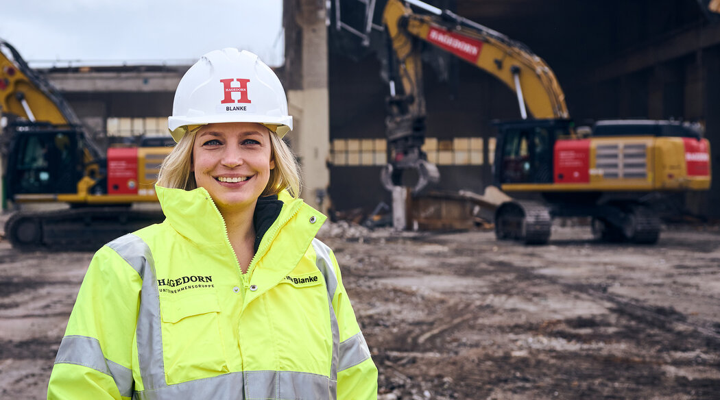 Blonde Frau mit Helm und gelber Sicherheitsjacke. Im Hintergrund ist ein Bagger zu sehen.